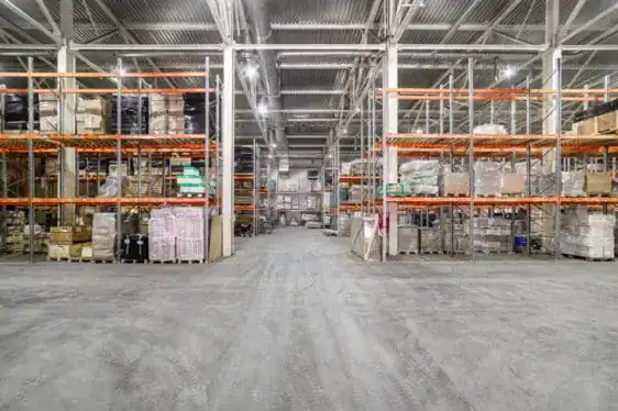 A large, well-lit warehouse with high shelving units filled with boxes and palletized goods. A wide aisle runs through the center.