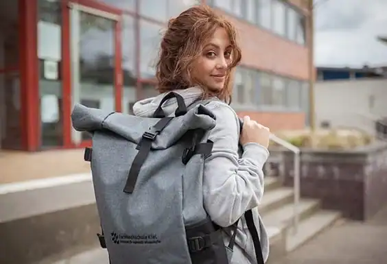 Junge Frau mit rötlich-braunen Haaren und hellgrauem Kapuzenpullover, die einen grauen Rucksack trägt und über die Schulter blickt.