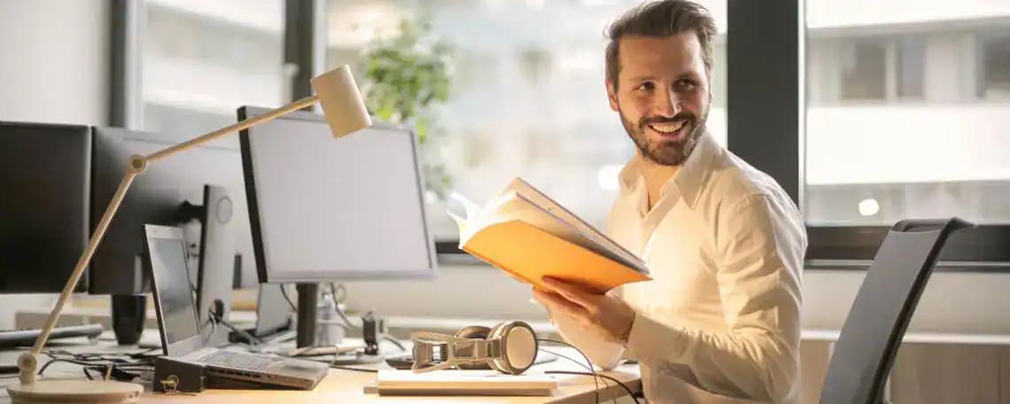 Ein lächelnder Mann mit Bart und weißem Hemd sitzt an einem Schreibtisch mit zwei Monitoren, Laptop und Schreibtischlampe. Er hält ein orangefarbenes Notizbuch in den Händen und blickt zur Seite.