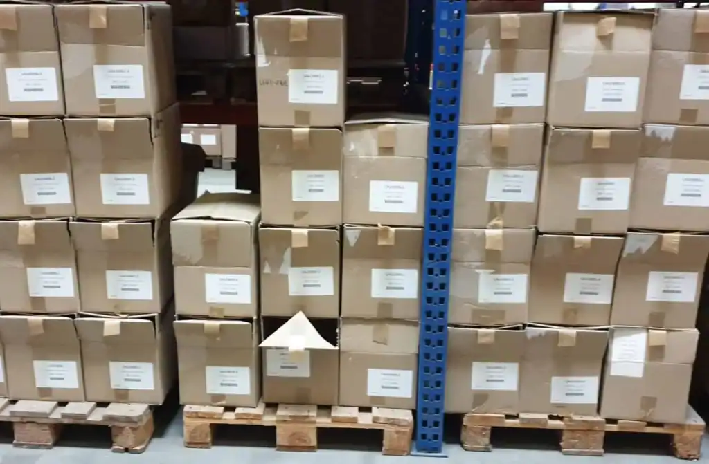 Stacked cardboard boxes on wooden pallets in a warehouse, with white labels and tape, blue shelving in the foreground.