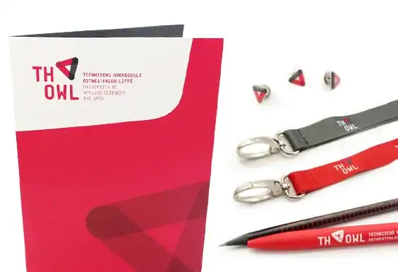Merchandising-Artikel der TH OWL: roter Ordner, Anstecker, graues und rotes Schlüsselband, Bleistift, roter Kugelschreiber.
