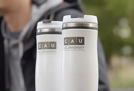 Zwei weiße Thermobecher mit dem Logo der CAU Kiel University stehen nebeneinander, eine Person ist unscharf im Hintergrund.